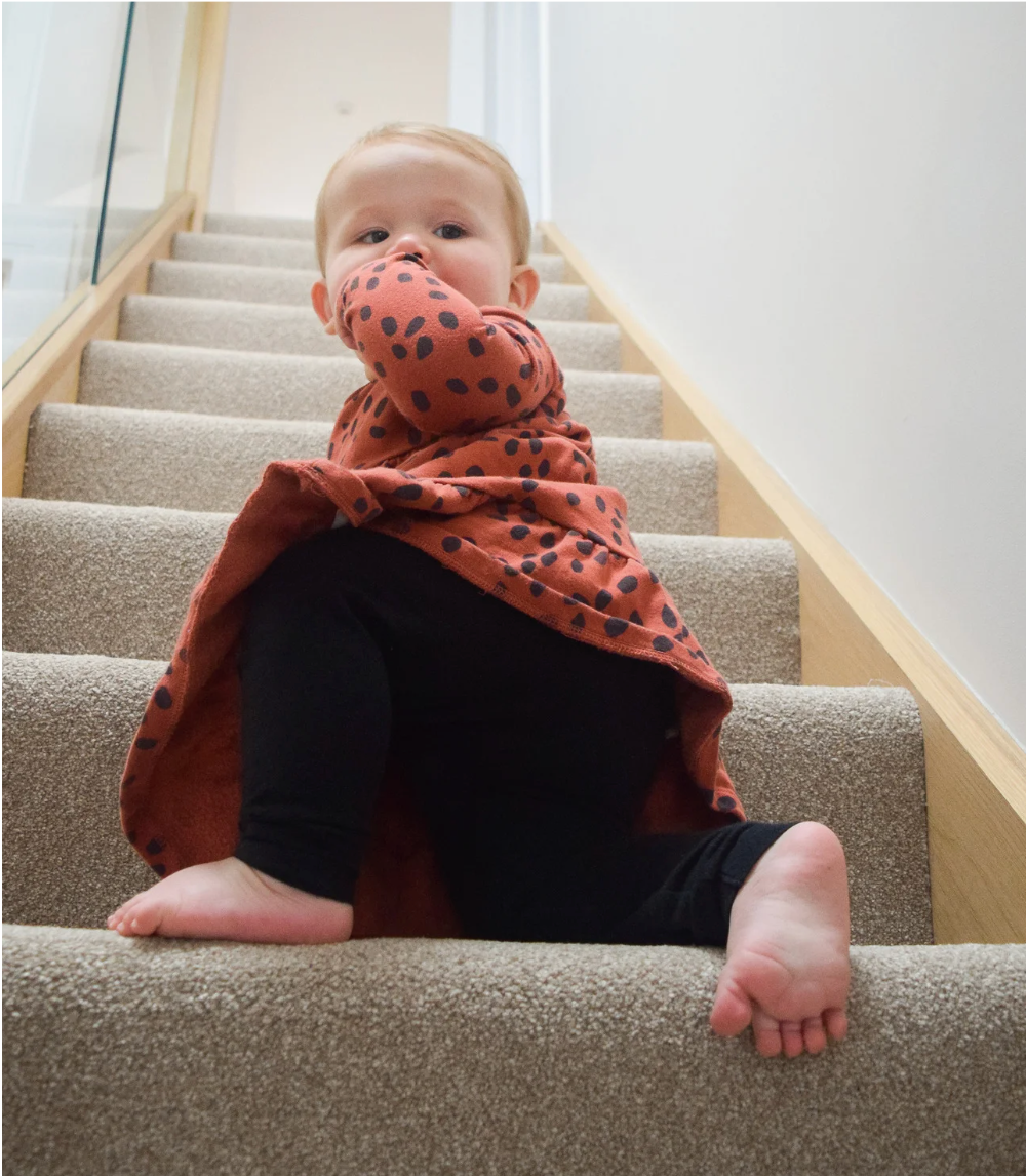 A toddler in a rust-colored dress with black spots and black leggings climbs up a carpeted staircase, looking up with one hand in their mouth.