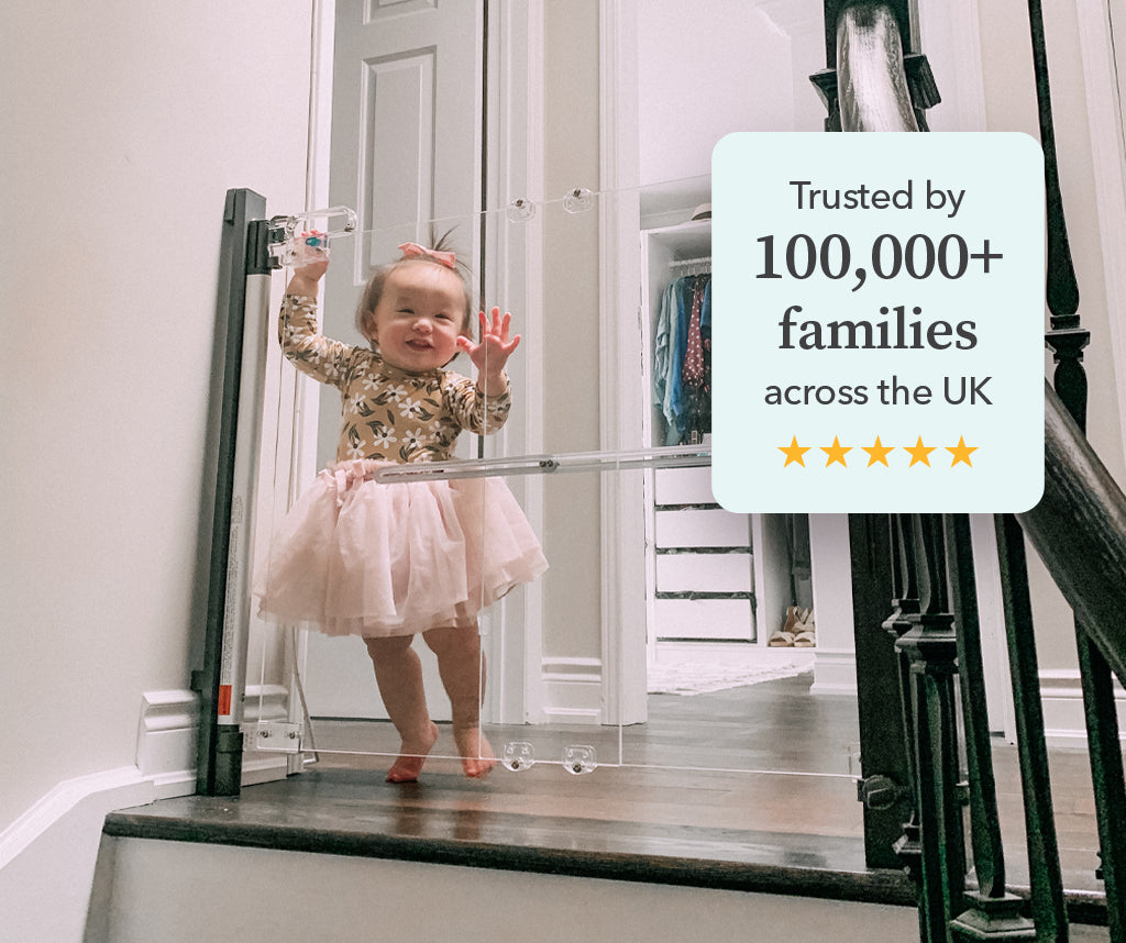 A toddler in a pink tutu stands against a clear pressure fitted stairgate. The image has a text overlay reading 