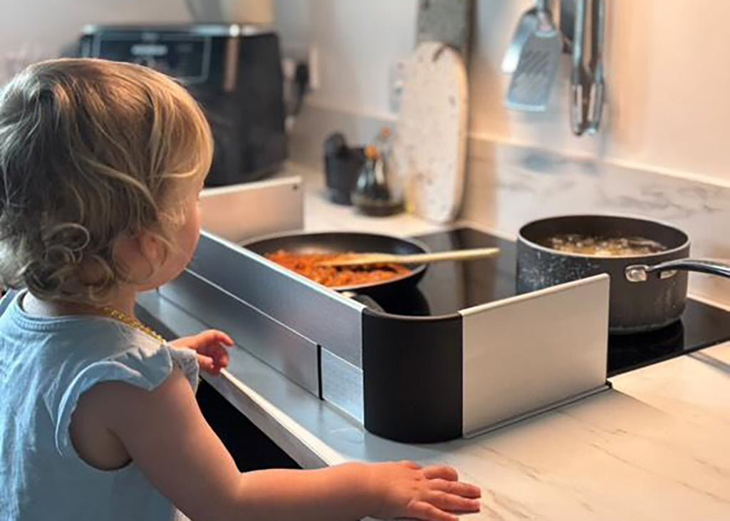 A toddler in a light blue shirt stands at a modern black and stainless steel stovetop, watching food cook in pans on a white marble kitchen countertop.