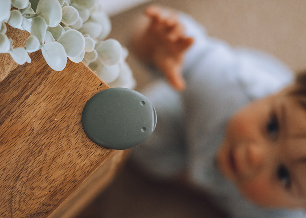 A close-up of a round, matte grey corner safety guard installed on a wooden table corner, with a baby in a light blue outfit sitting nearby and white flowers in the top-left corner.