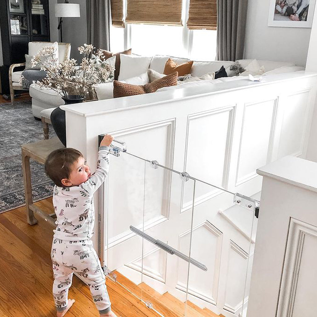 A toddler in a white animal-print pajama set stands on a wooden staircase landing and pushes a closed stair gate with clear panels. The scene is set in a bright, modern living room with a white sofa and a patterned rug.