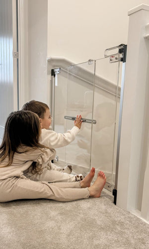 A toddler and a young girl sit on a carpeted floor in a home setting, looking at and touching a clear glass stair gate with a silver metal handle and black hardware.